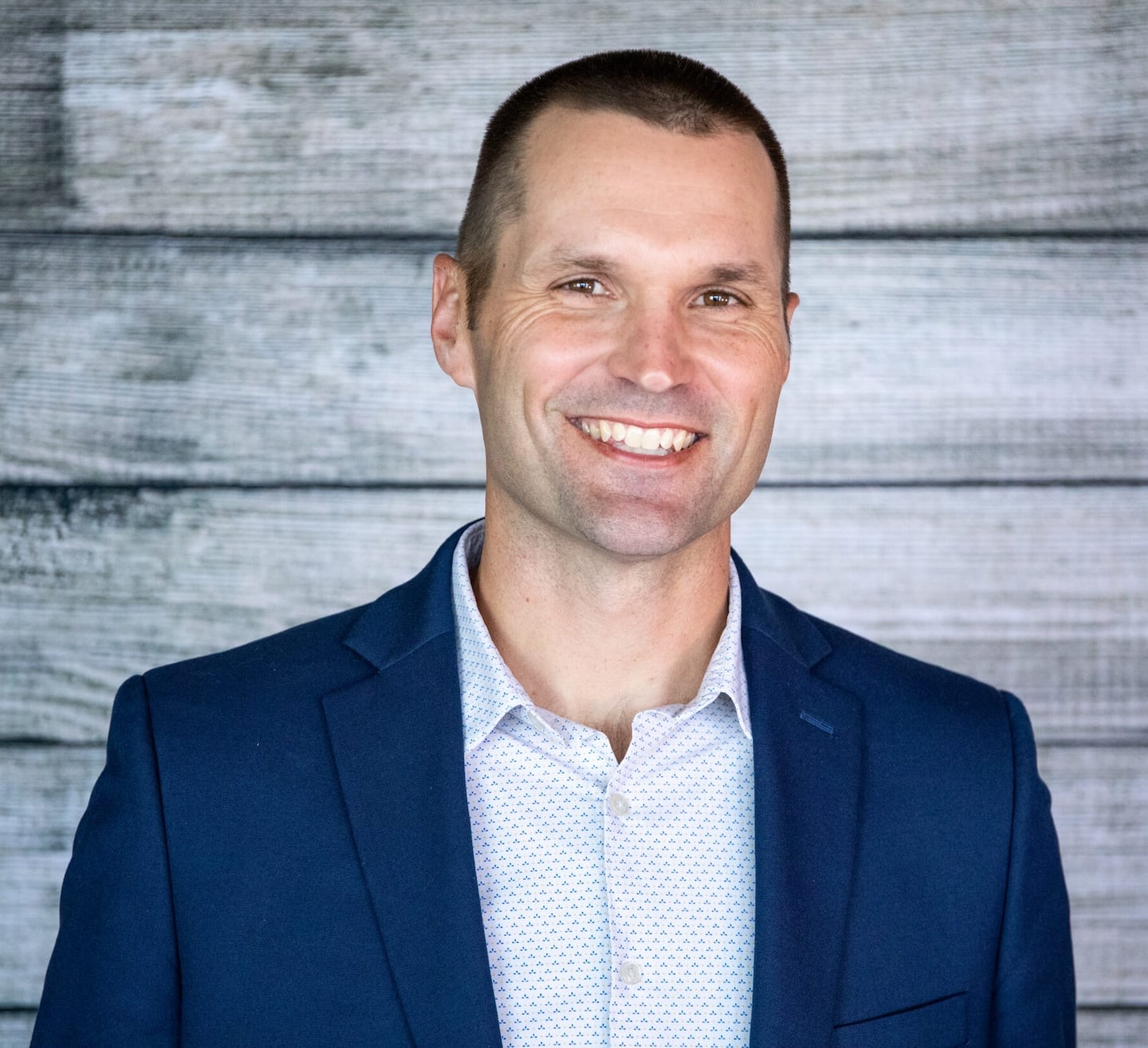 Marcus Sheridan, in a blue blazer and white patterned shirt, confidently smiles before a wooden plank background on Hook Agency's site.